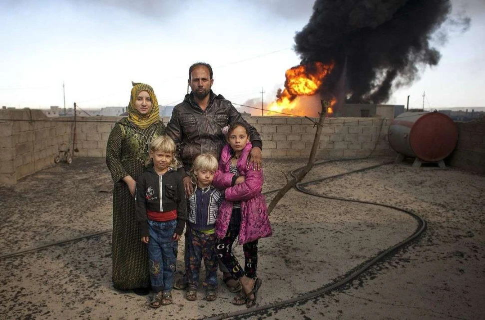 Family of five stands on a rooftop near a burning building. Woman in headscarf, man in jacket, and three children look forward.