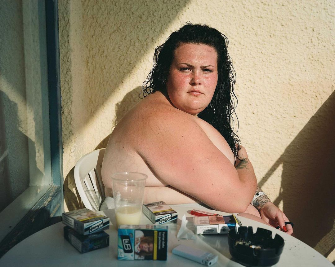 Woman with dark curly hair sits at a table with cigarettes and a drink, creating a candid, unfiltered lifestyle portrait.