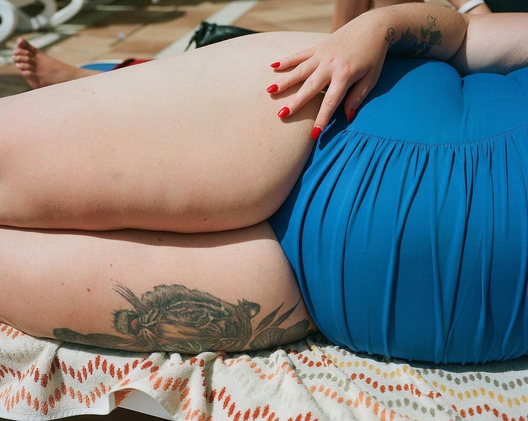 Woman in blue swimsuit lying on patterned towel in bright sunlight, showing tattoos and red nails. Relaxed, summery scene.