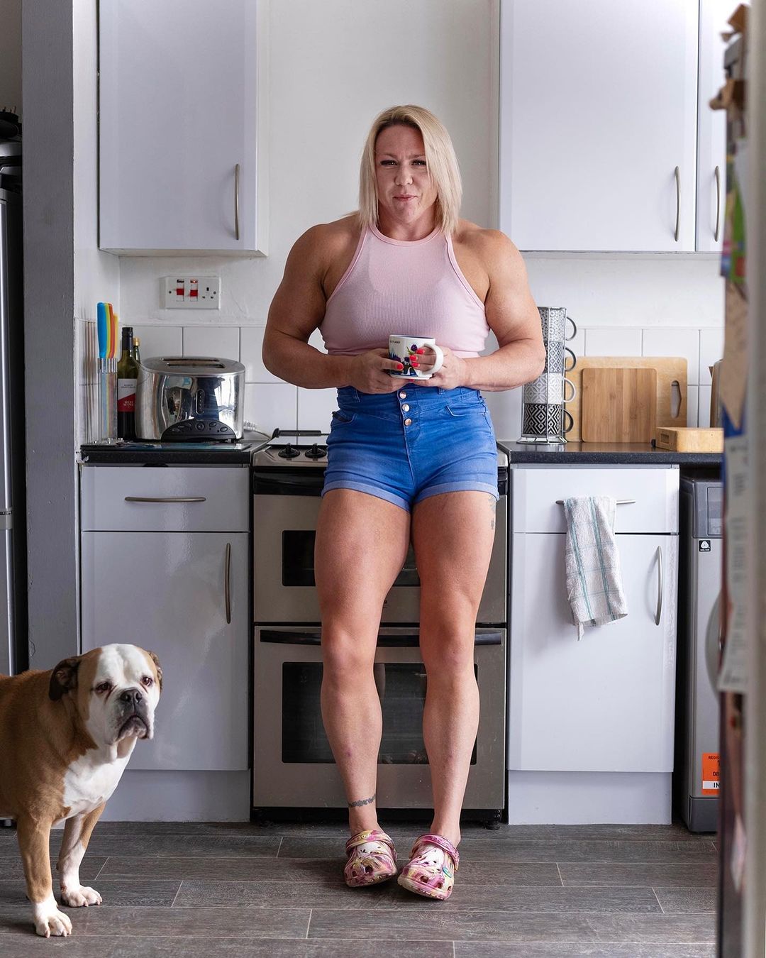 A muscular woman in denim shorts poses in a bright kitchen while holding a mug, standing next to a brown and white bulldog.