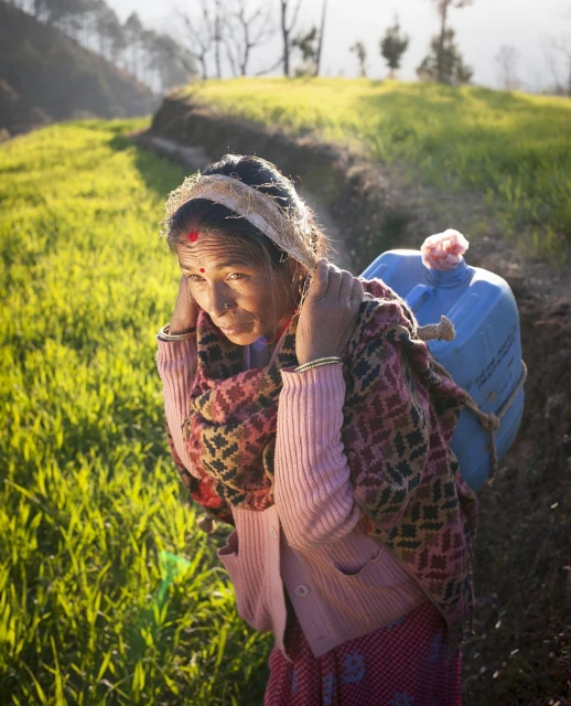 An elderly woman carries a blue jug over her shoulders while walking through a sunlit grassy field in a rural environment.