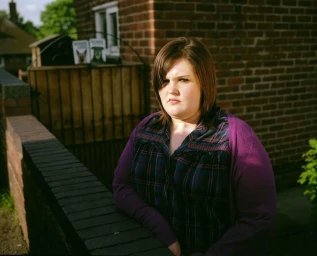 Young woman standing outdoors near a brick wall wearing a dark plaid shirt and purple cardigan