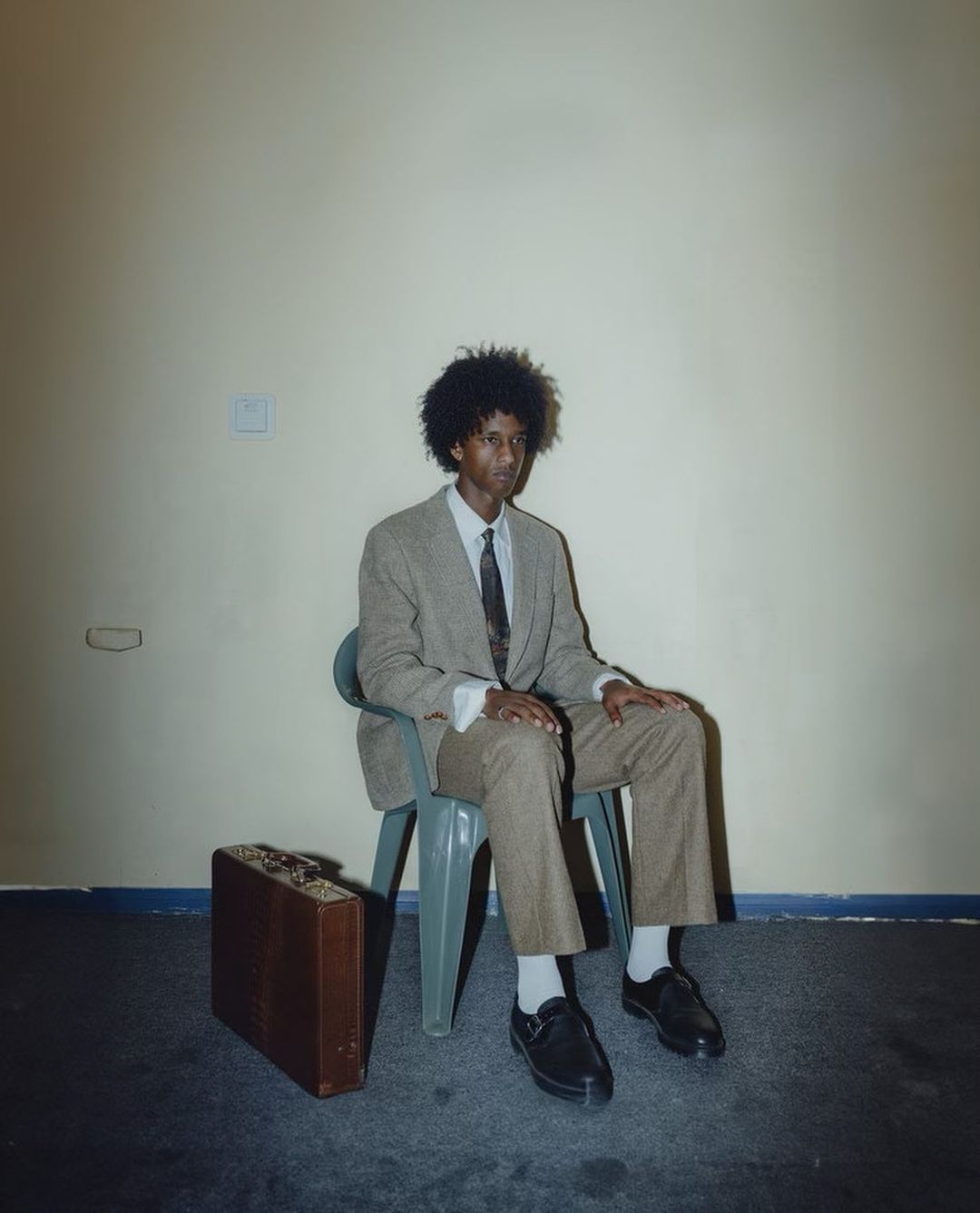 Portrait of an adult male sitting on a plastic chair in suit and tie with a briefcase next to him, photographed in a room with a light wall.