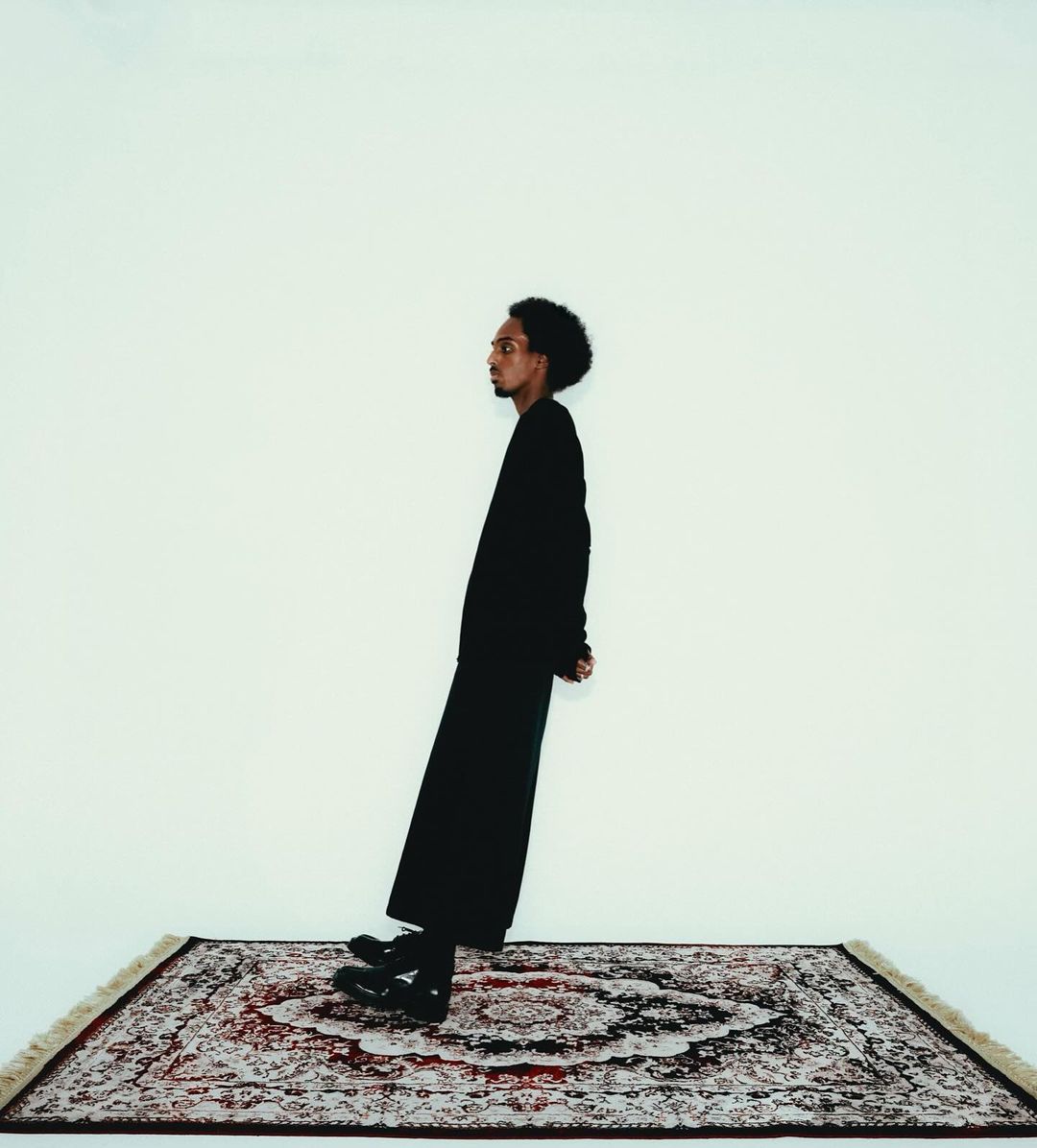 A man with dark hair stands at an angle on an oriental rug against a white backdrop in a studio-style portrait shot. He wears all black.