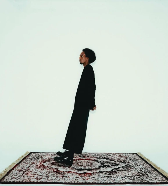 A man with dark hair stands at an angle on an oriental rug against a white backdrop in a studio-style portrait shot. He wears all black.