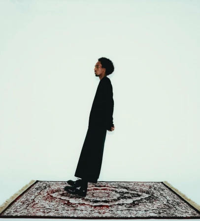 A man with dark hair stands at an angle on an oriental rug against a white backdrop in a studio-style portrait shot. He wears all black.