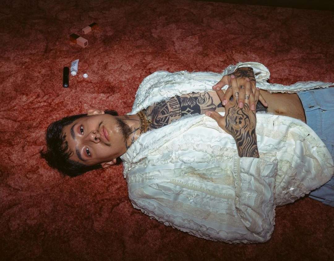 Young adult male with tattoos lies on a red carpet in a white lace jacket with beauty products nearby in an indoor setting.