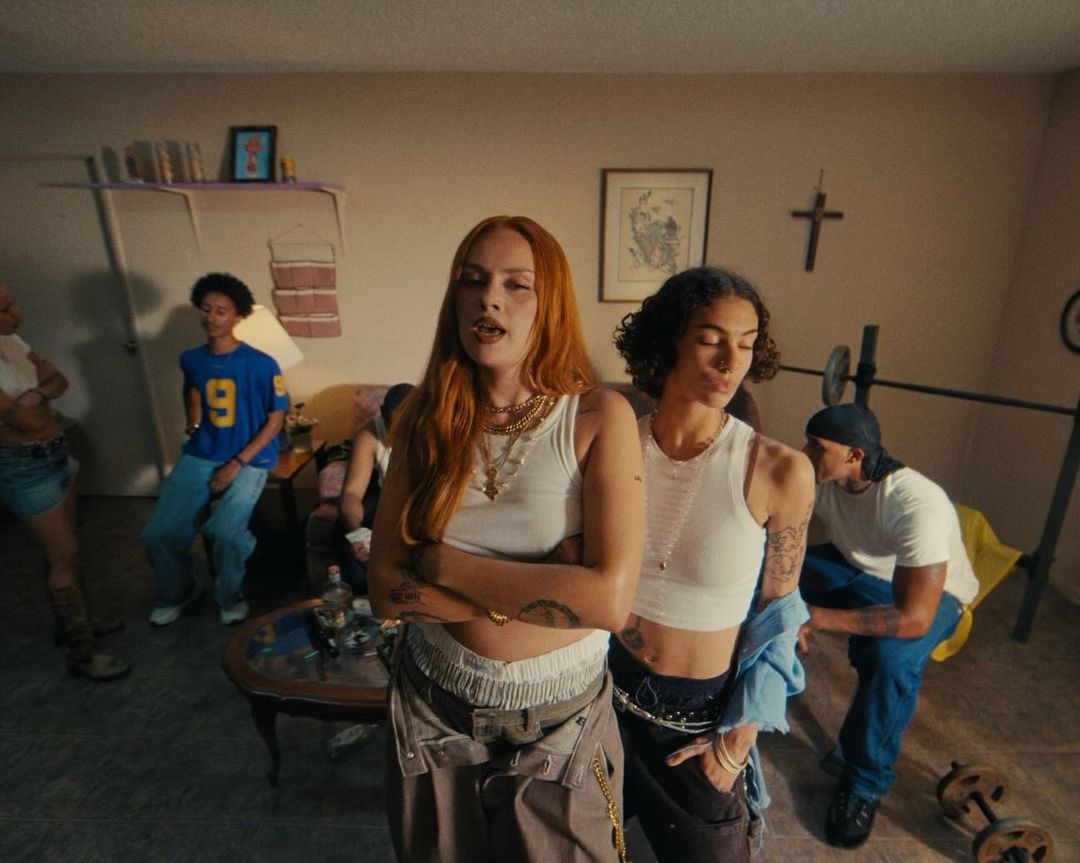 Group of young adults casually posed in a living room, featuring two women in crop tops and baggy pants in the foreground.