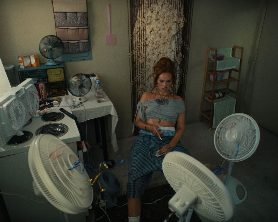 Adult female sitting with multiple fans in an interior setting with walls, a stove, a table, and other household objects and furnishings.