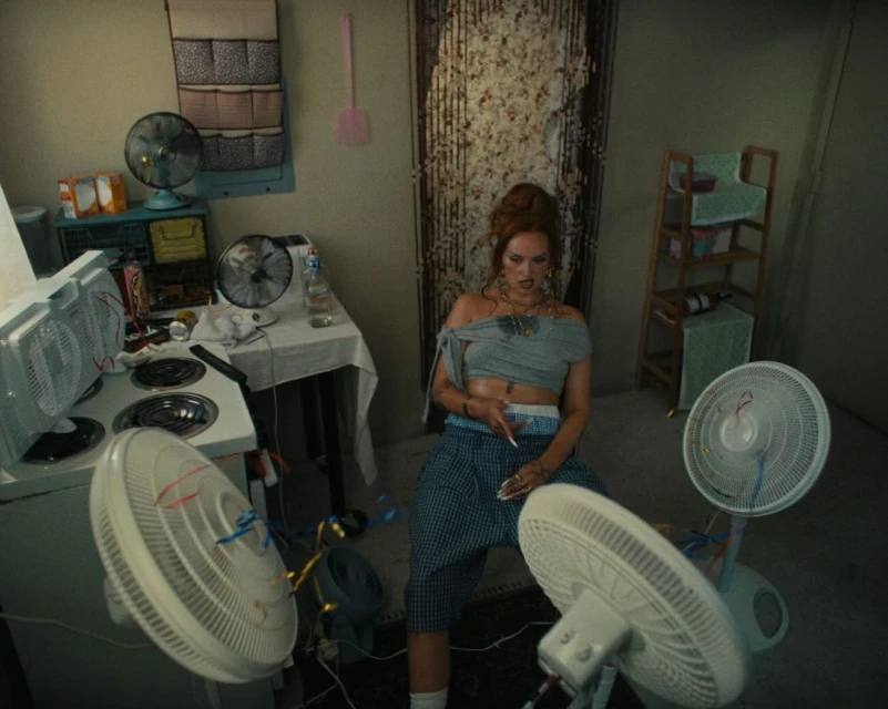 Adult female sitting with multiple fans in an interior setting with walls, a stove, a table, and other household objects and furnishings.