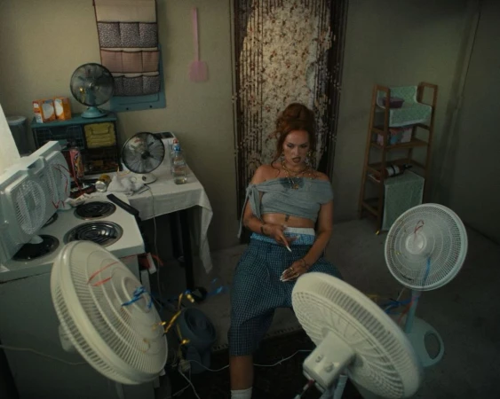Adult female sitting with multiple fans in an interior setting with walls, a stove, a table, and other household objects and furnishings.