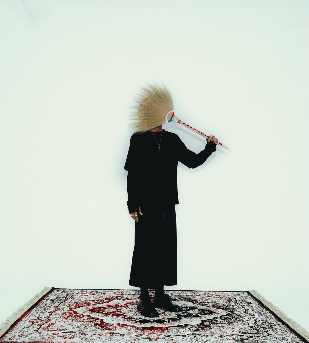Person in dark clothing poses on an ornate rug in front of a white wall, obscuring their face with a decorative object.