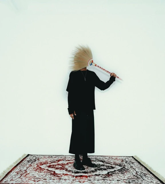Person in dark clothing poses on an ornate rug in front of a white wall, obscuring their face with a decorative object.