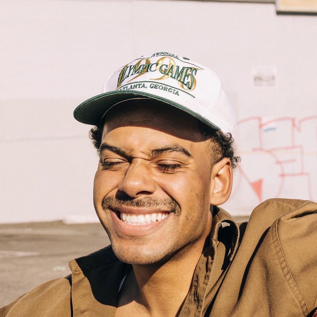 Portrait of a young adult man wearing an Olympic Games baseball cap, smiling with eyes closed, and a brown collared shirt outdoors.