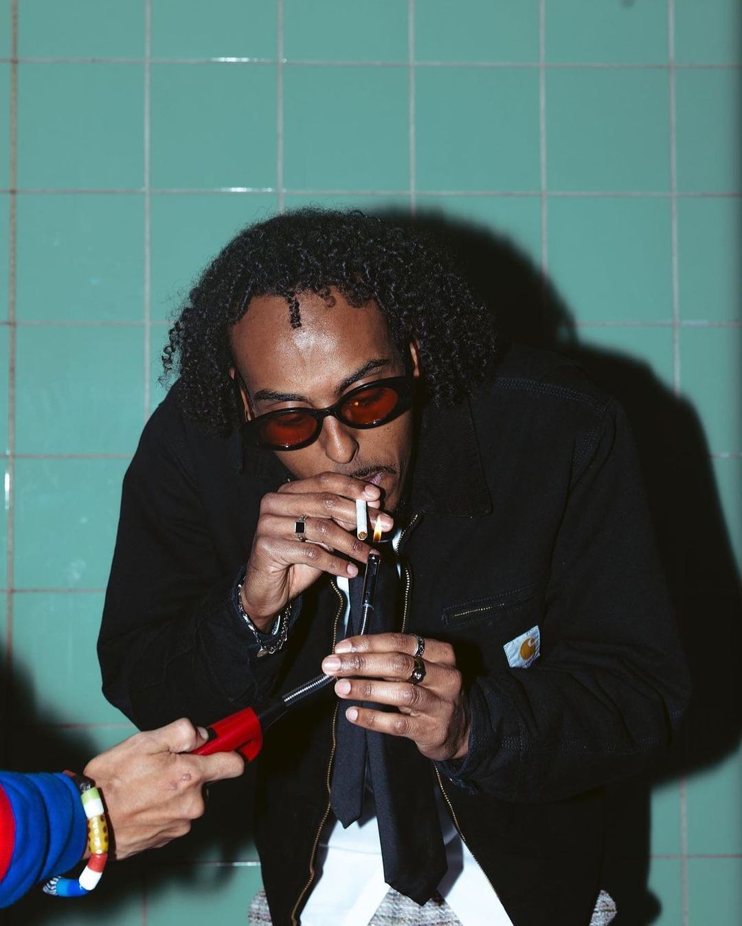Stylish man with curly hair and sunglasses smokes a cigarette indoors against a tiled wall, illuminated by artificial lighting.