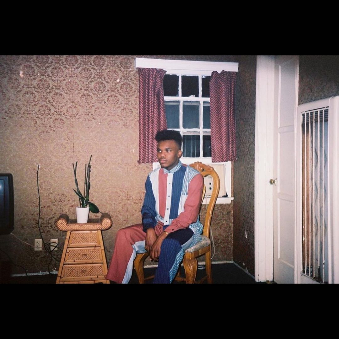 An adult man in a color-blocked suit sits in a vintage-style room with patterned wallpaper and a plant stand, under the light of a window.