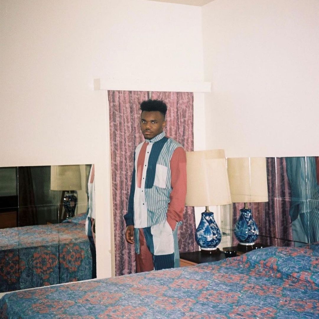 A stylish African-American man in patchwork shirt and pants poses in an indoor setting with a bed, lamps, and white walls.