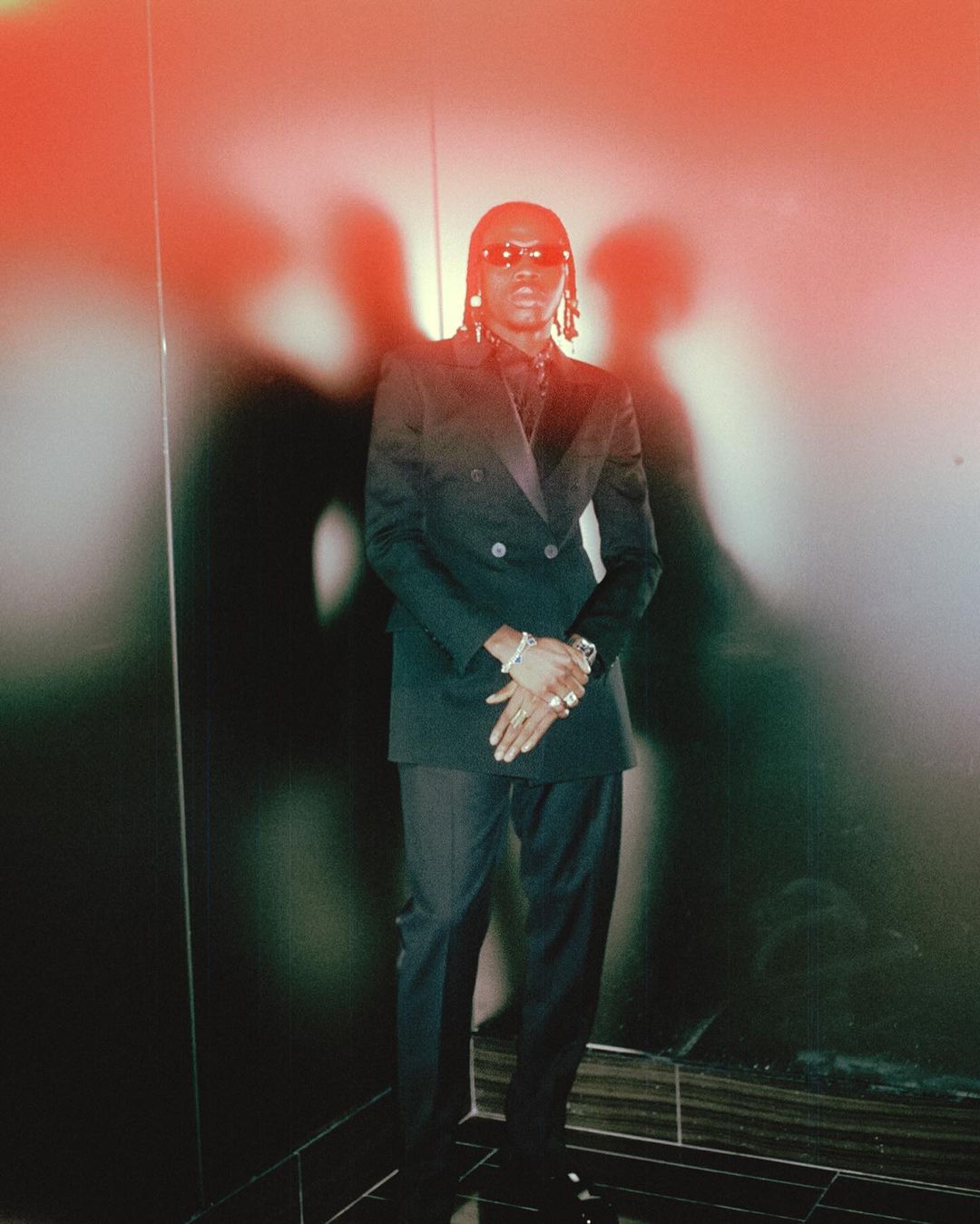 Man wearing a dark double-breasted suit and sunglasses standing against a wall with strong shadows in New York.