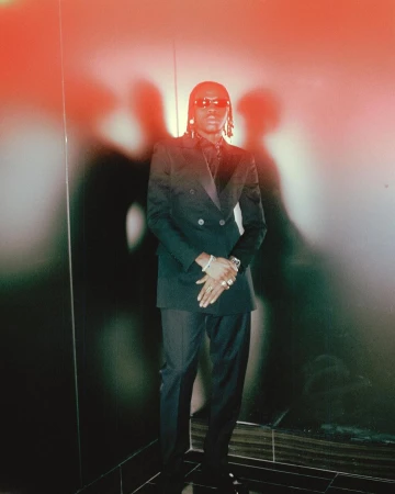 Man wearing a dark double-breasted suit and sunglasses standing against a wall with strong shadows in New York.