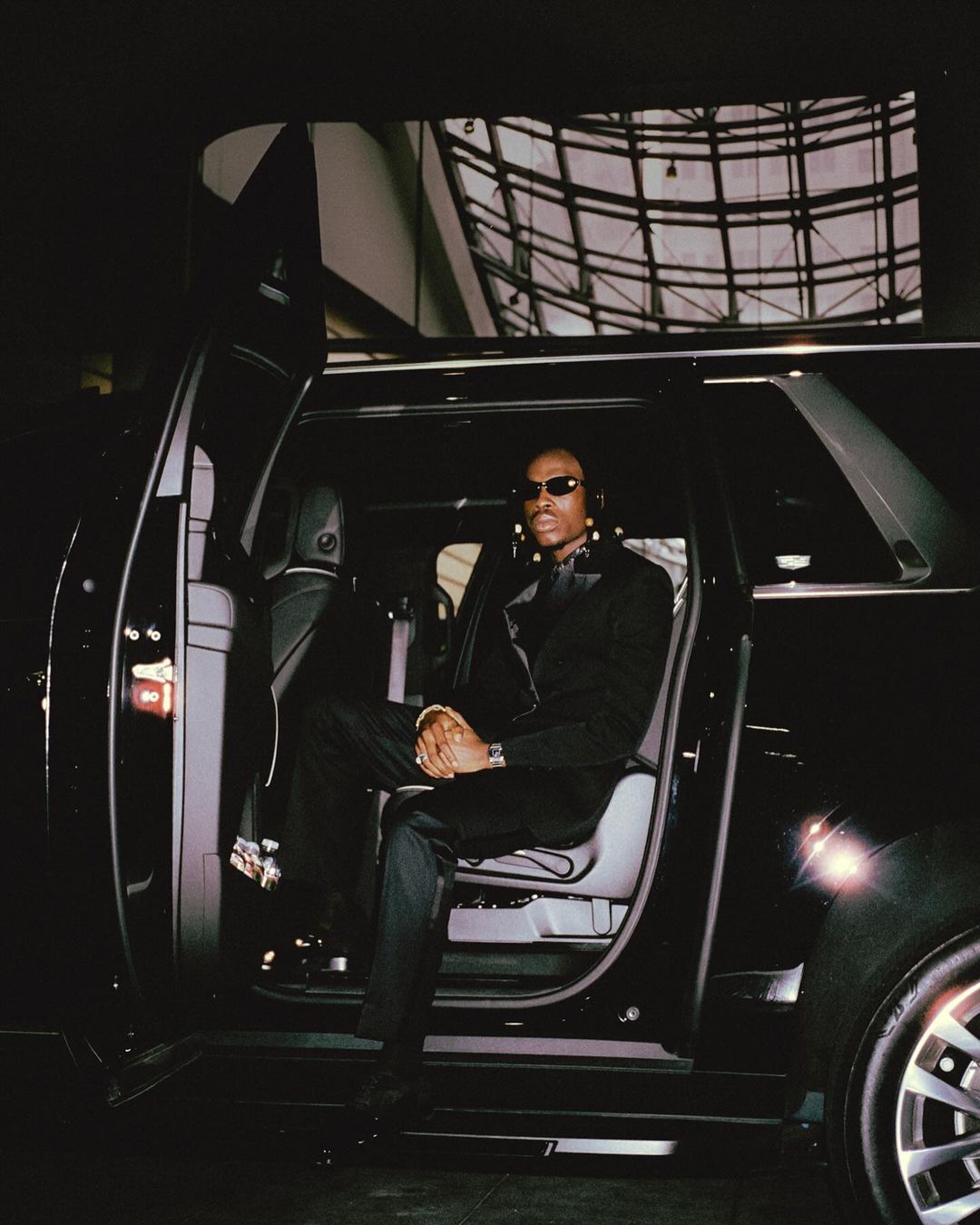 Man in sunglasses and formal wear exits a black SUV with a modern building in the background, capturing a stylish and upscale moment.