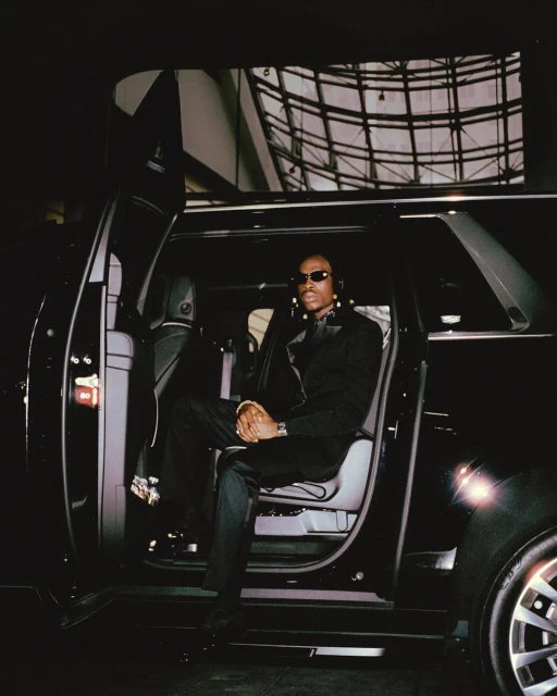 Man in sunglasses and formal wear exits a black SUV with a modern building in the background, capturing a stylish and upscale moment.