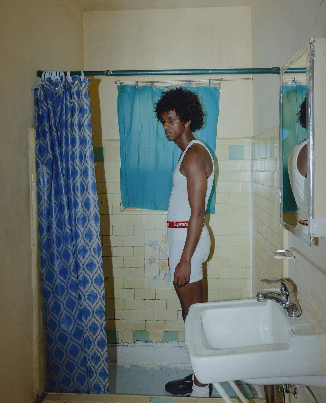 Young man in tank top and underwear stands in a bathroom, with blue patterned shower curtain, sink, and tiled walls.