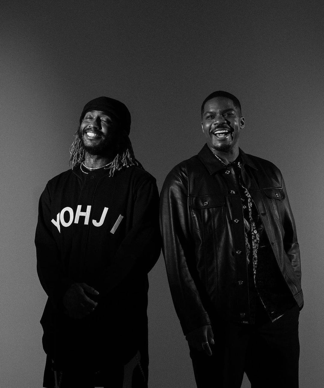 Two adult men in a monochrome portrait; one with dreadlocks and a beanie, the other in a leather jacket, both smiling against a gray backdrop.
