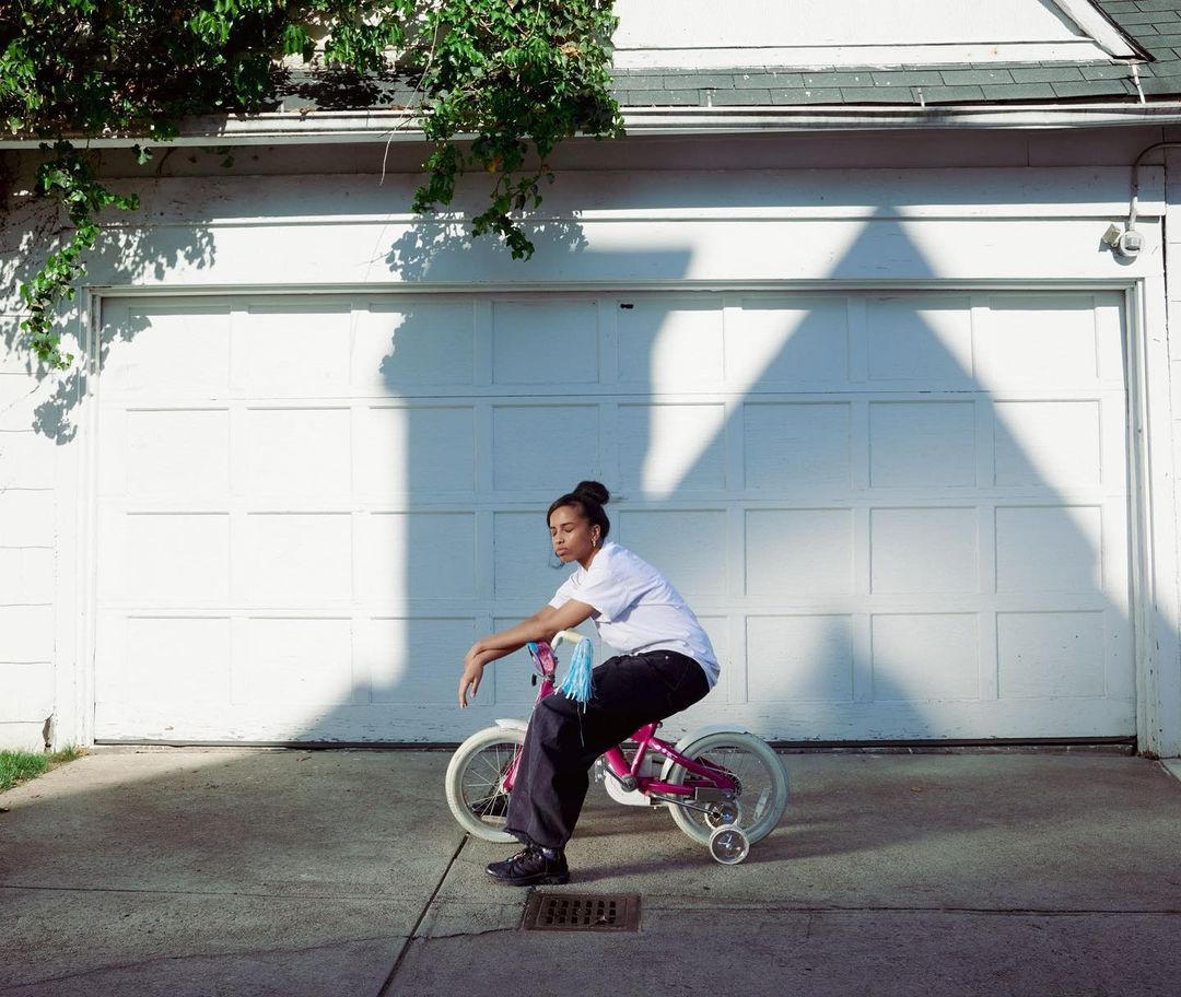 Young adult rides a small pink bicycle with training wheels in front of a white garage door, bathed in sunlight and shadows.
