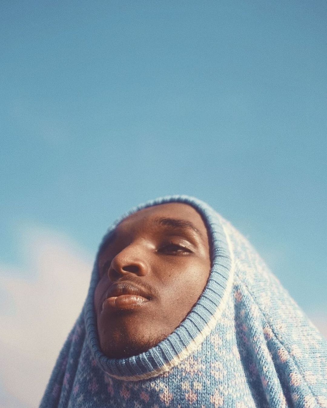 Portrait of an adult man with dark skin wearing a patterned blue and white knit balaclava against a bright blue sky background.