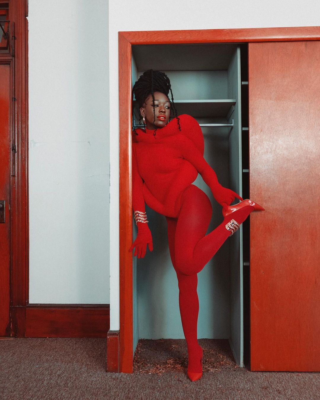 A woman in a red bodysuit and matching heels poses in a doorway with red trim, her leg raised, in a brightly lit indoor setting.
