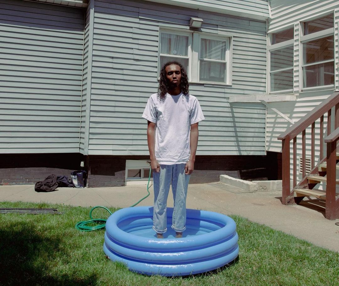 Person standing in blue inflatable swimming pool on lawn in backyard on summer day in front of house with siding and windows.