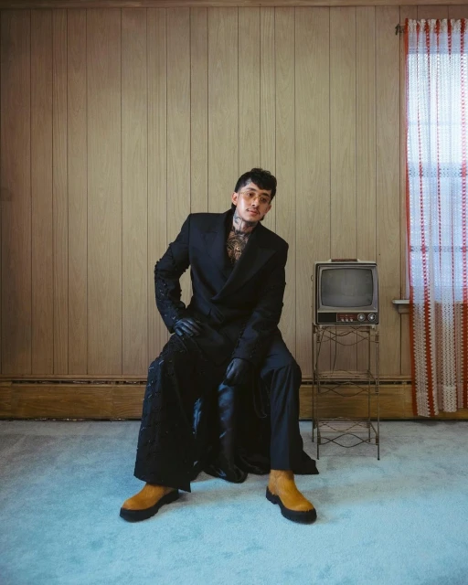 Stylish man with tattoos poses in a black suit next to a vintage television in a wood-paneled room with light blue carpet.