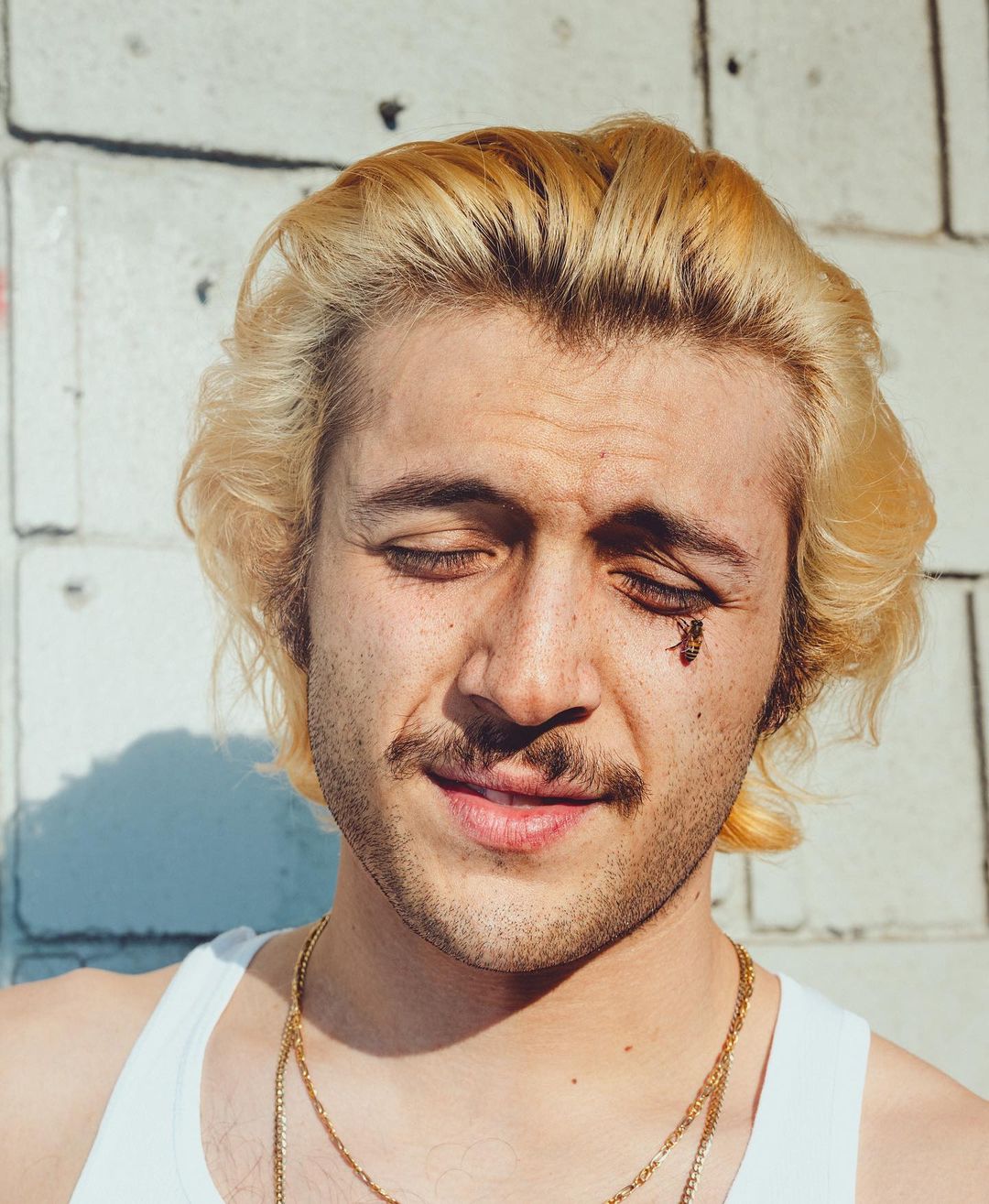 Close-up portrait of a young adult male with blond hair and a mustache. He is wearing a white tank top and gold chain necklaces.