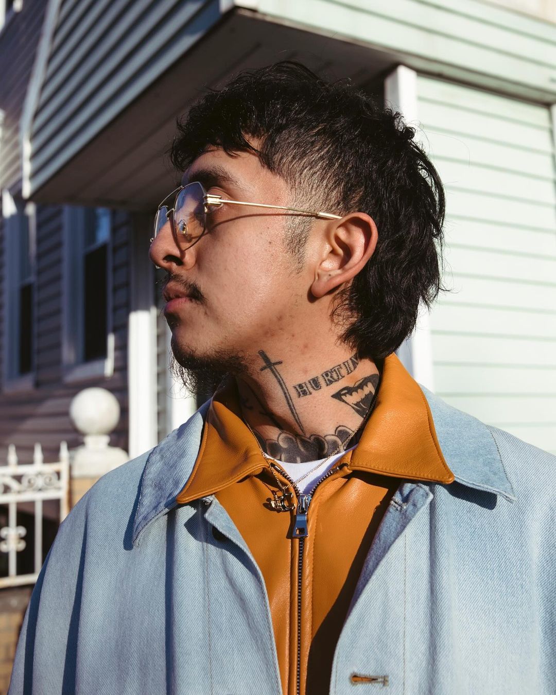 Portrait of a young man with a mullet hairstyle and neck tattoos wearing glasses and a jean jacket standing outside a house.