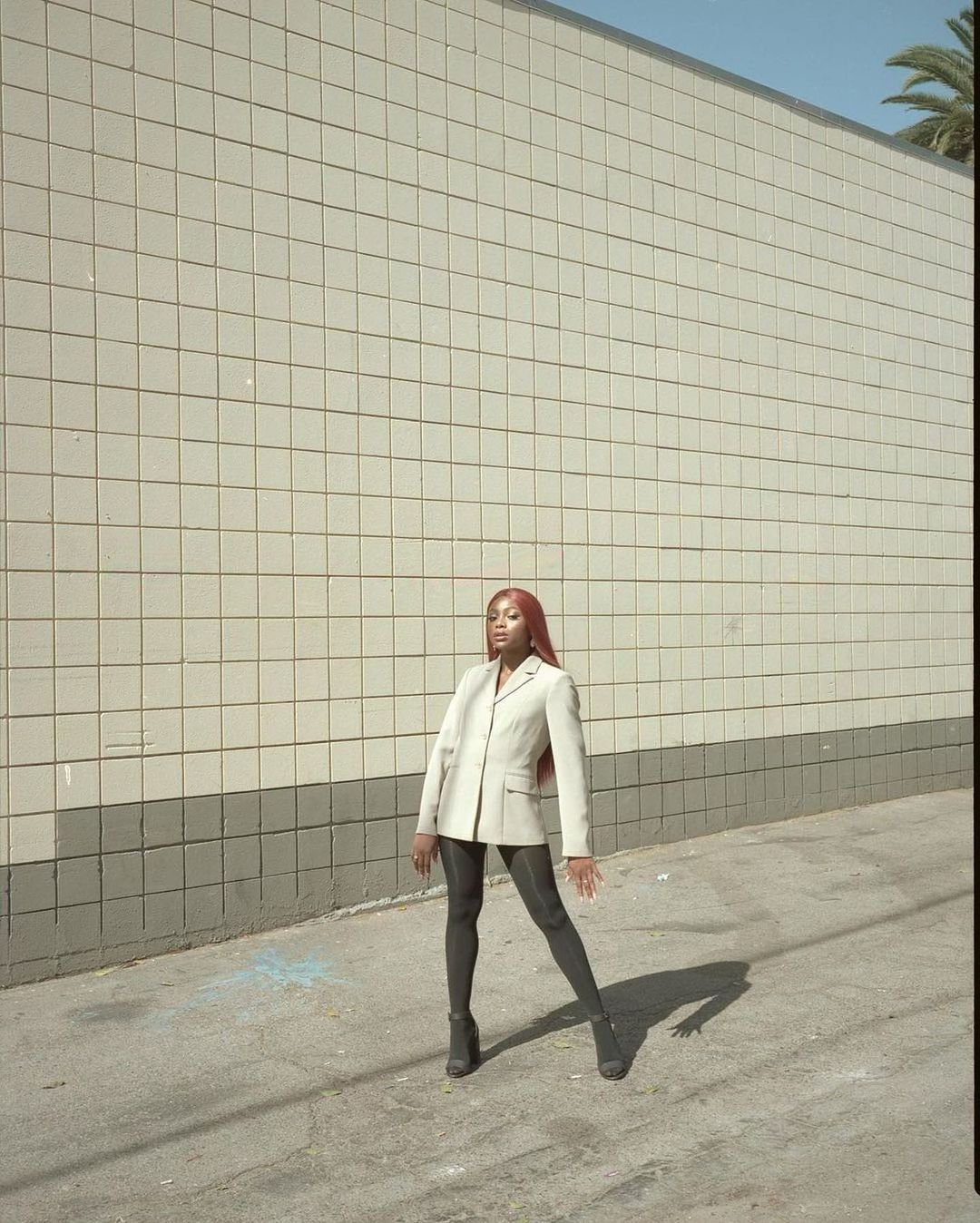 An adult woman with red hair is standing on a concrete surface in front of a tiled wall, wearing a business jacket, leggings and high heels.