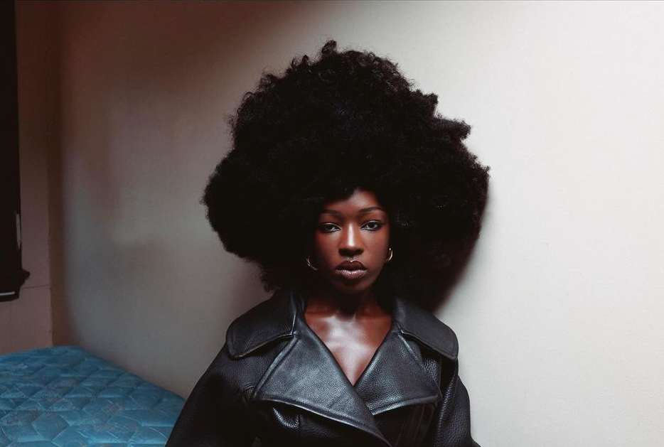 Indoor portrait of a Black woman with a large afro hairstyle, wearing a black leather coat, standing in front of a simple wall.