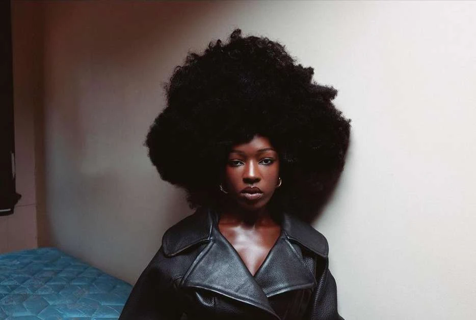 Indoor portrait of a Black woman with a large afro hairstyle, wearing a black leather coat, standing in front of a simple wall.