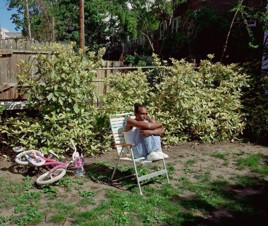 Young adult sits on a lawn chair in a grassy backyard next to a child's pink bicycle; plants, a fence, and a brick building are visible.