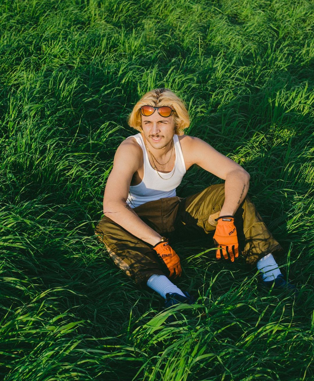 A young adult male with blonde hair sits cross-legged in a bright green field wearing sunglasses, a white tank top, and work gloves.