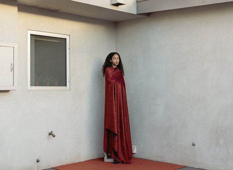 Young woman in a red wrap stands in an outdoor corner with a white window. Simple fashion portrait with blank concrete wall.