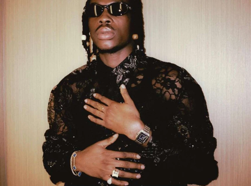 Adult male poses in a black lace shirt and sunglasses with jewelry against a neutral wall, captured in a medium shot.