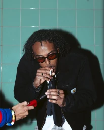 Man with curly hair and sunglasses lighting a cigarette with a red lighter against green tiles, photographed by Abdi Ibrahim