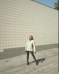 Woman in a light beige blazer and black tights posing against a light tiled outdoor wall under sunlight