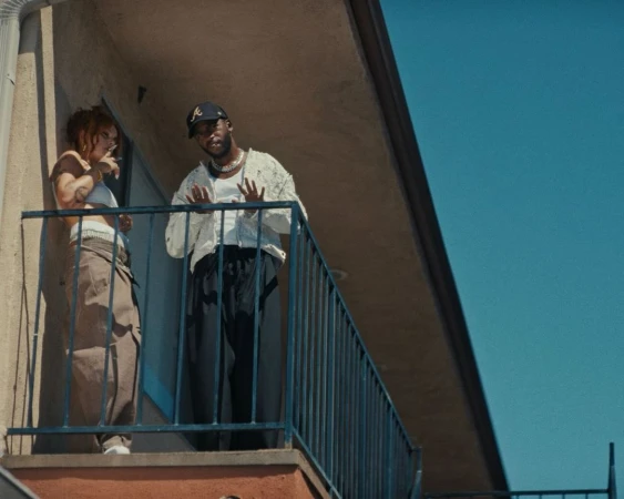 A man in a baseball cap and white jacket standing next to a woman leaning on a balcony railing against a blue sky.