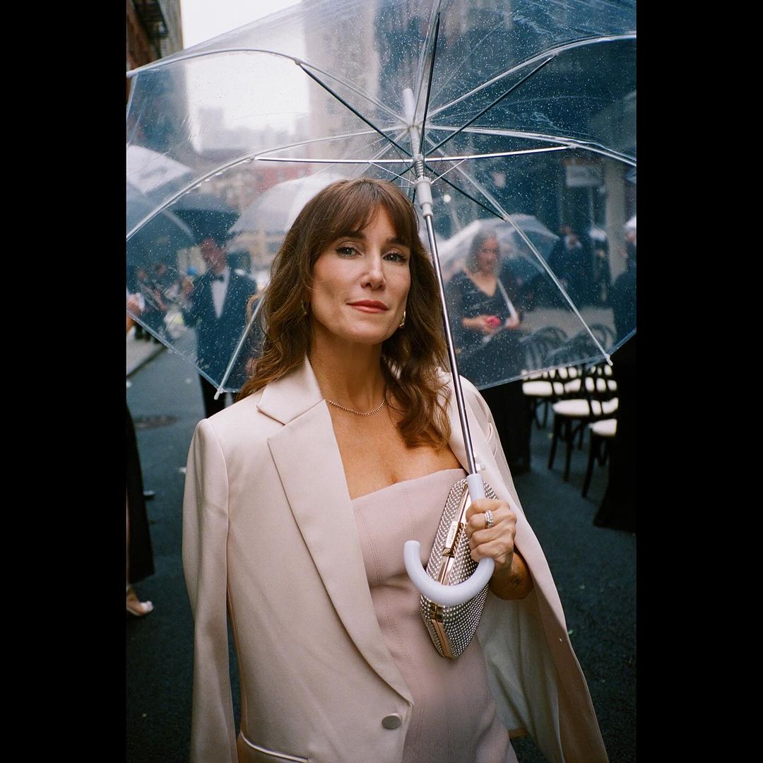A woman stands in the rain under a clear umbrella wearing a light pink suit with a building and people in the background.