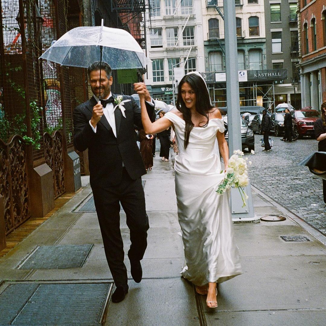 Couple walking on a city sidewalk in the rain under an umbrella. The woman is in a wedding dress holding flowers.
