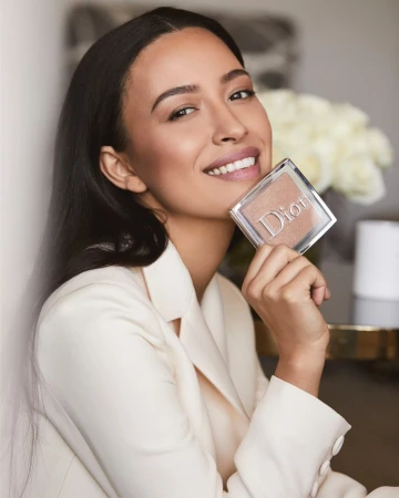 A woman smiles holding Dior makeup compact. She wears a white suit. Beauty shot featuring makeup product.