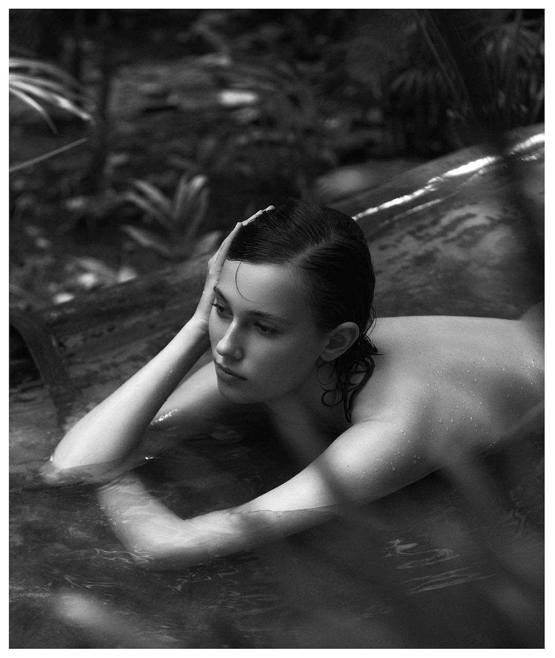 Black and white portrait of a fair-skinned woman in water with palm fronds, looking away with hand on her head in a thoughtful pose.