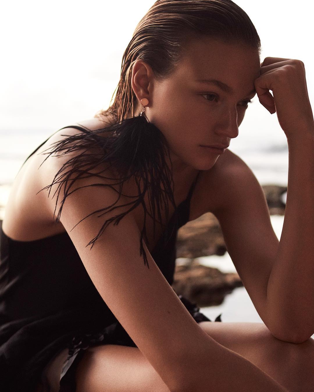 Adult woman with slicked-back hair sits outside wearing a black dress and a feather earring with neutral tones and soft lighting.