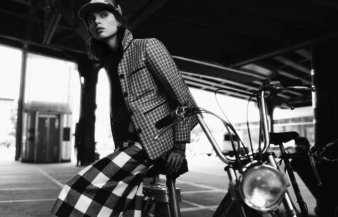Black and white portrait of a stylish adult woman posing on a vintage motorcycle, wearing a plaid blazer and pants in an urban setting.